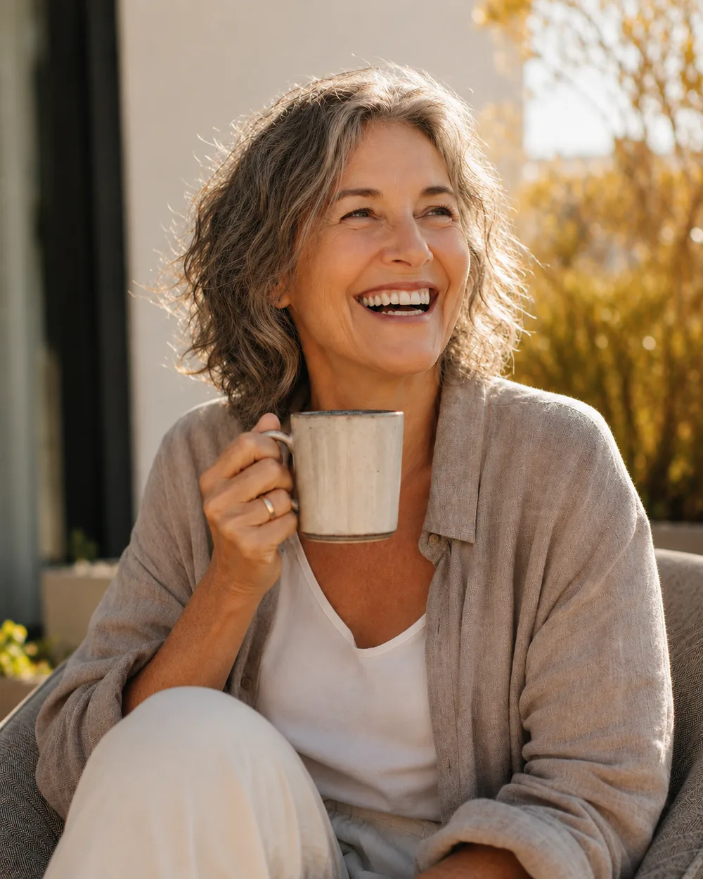 A woman in her late fifties laughing over a mug of coffee in the morning sun